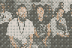 Thomas in a white shirt with blue jeans, next to Yoshie in a black shirt with lots of cat graphics and blue pants, sitting in the midst of the KirbyKonf crowd, smiling, listening to a talk.