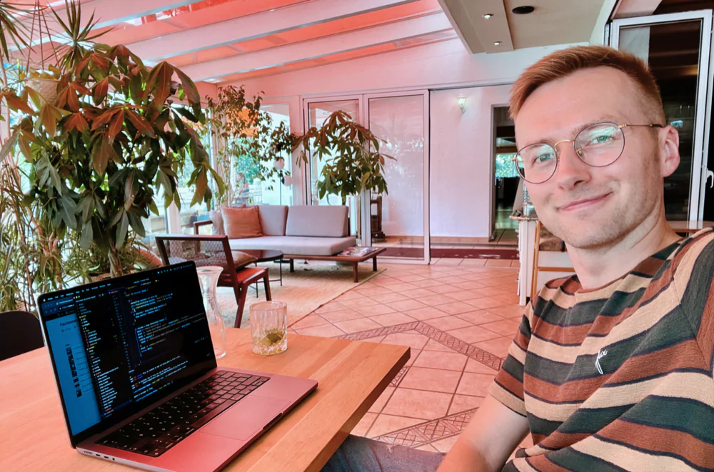 A man sitting in a cafe, in front of a laptop, smiling at the camera. The laptop's screen shows code.