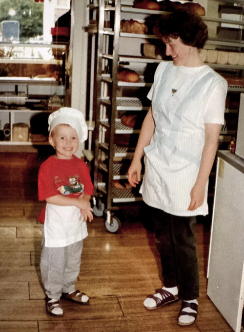 A little boy wearing a baker's hat and apron, standing in the back of his parents' bakery, smiling next to his mother.