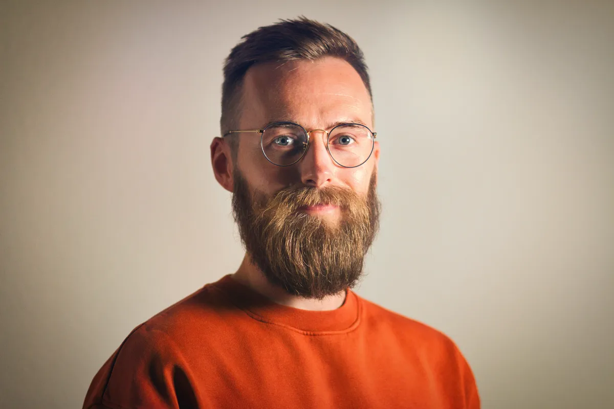 Close-up portrait of a bearded Thomas in his thirties, wearing round glasses and an orange sweater.
